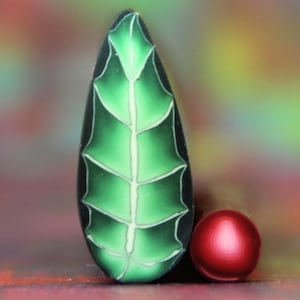 Op de afbeelding: Een groen, bladvormig ornament met witte aderen en een kleine rode bol. Het blad heeft een glanzende afwerking en een donkergroene omtrek. De rode bol is glad en rond. De achtergrond is een vage mix van kleuren.