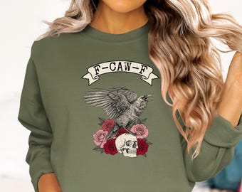 May include: Olive green sweatshirt with a graphic featuring a raven with outstretched wings, perched atop a skull and roses. The banner above the raven reads "F-CAW-F". The design incorporates red, pink, and white elements.