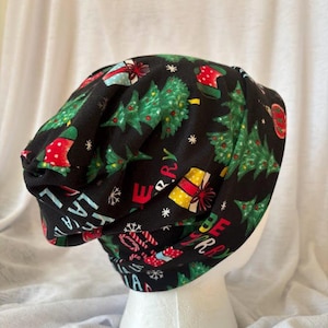 Op de afbeelding: Zwarte beanie-muts met een kerstpatroon. De muts heeft groene kerstbomen, rood en geel verpakte cadeaus en de woorden "Merry Christmas" in het wit. De hoed wordt getoond op een witte mannequinhoofd.