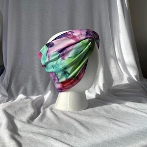 May include: A vibrant floral headband in pink, purple, and green hues. The fabric is gathered and knotted, forming a fashionable accessory. The headband is presented on a white mannequin head, against a white backdrop.