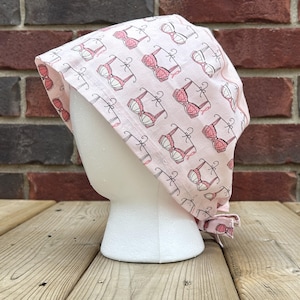 May include: A light pink surgical scrub cap with a repeating pattern of pink and white bras. The cap is tied at the back and is displayed on a white mannequin head. The background includes a brick wall and wooden planks.
