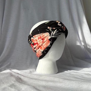 May include: A black floral headband with coral and white flower details. The headband is displayed on a white mannequin head. The background is white.