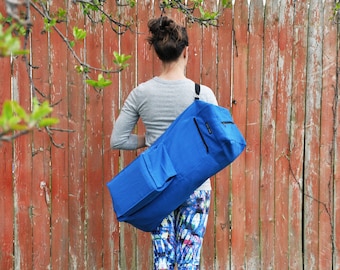 Puede incluir: Una persona llevando una bolsa azul para esterilla de yoga con una correa negra sobre el hombro. La bolsa tiene una cremallera y un bolsillo en la parte delantera.