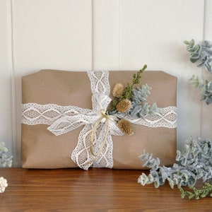 Pu&ograve; includere: Un pacchetto regalo in carta marrone legato con un nastro di pizzo bianco e decorato con un rametto di verde secco.