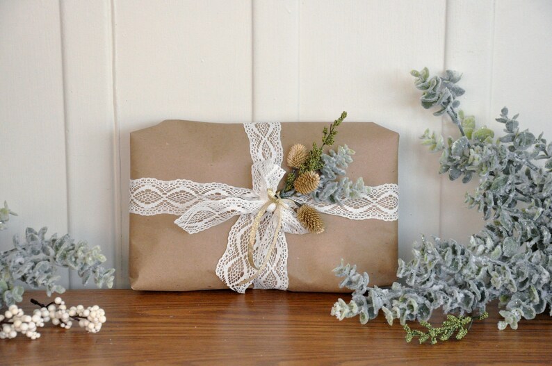 Pu&ograve; includere: Un pacchetto regalo in carta marrone legato con un nastro di pizzo bianco e decorato con un rametto di verde secco.