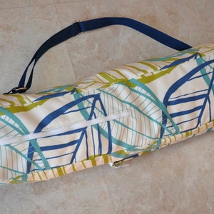 May include: A white yoga mat bag with a blue and green leaf pattern and a navy blue adjustable strap. The bag has a zipper closure.