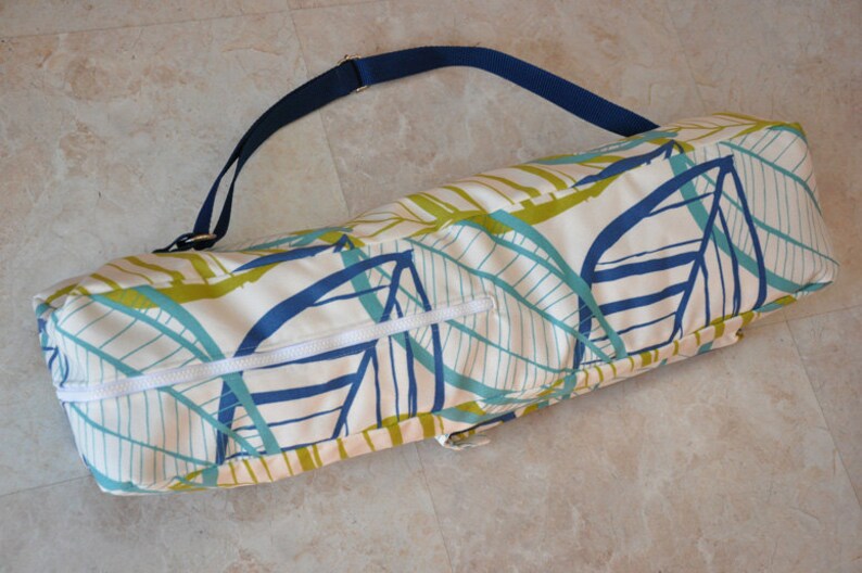 May include: A white yoga mat bag with a blue and green leaf pattern and a navy blue adjustable strap. The bag has a zipper closure.