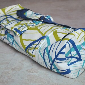 May include: A white yoga mat bag with a blue and green leaf pattern. The bag has a navy blue strap and a zipper closure.
