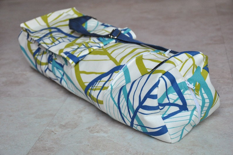 May include: A white yoga mat bag with a blue and green leaf pattern. The bag has a navy blue strap and a zipper closure.