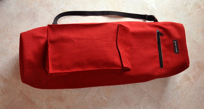 Pu&ograve; includere: Borsa per tappetino da yoga rossa con una tracolla nera e una tasca con cerniera. La borsa ha una piccola tasca sul davanti e il testo "Jenna & Co." stampato sul lato.