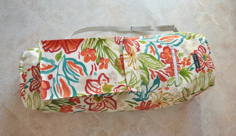 May include: A white canvas yoga mat bag with a floral print in shades of orange, red, green, and blue. The bag has a zippered pocket and a shoulder strap.
