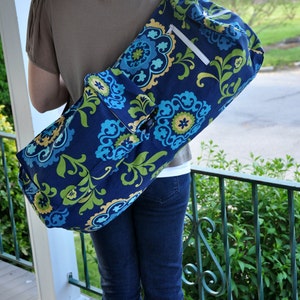 Pu&ograve; includere: Una donna porta una borsa per tappetino da yoga blu e verde con un motivo floreale e una tasca con cerniera. La borsa &egrave; indossata sulla spalla e ha una tracolla.