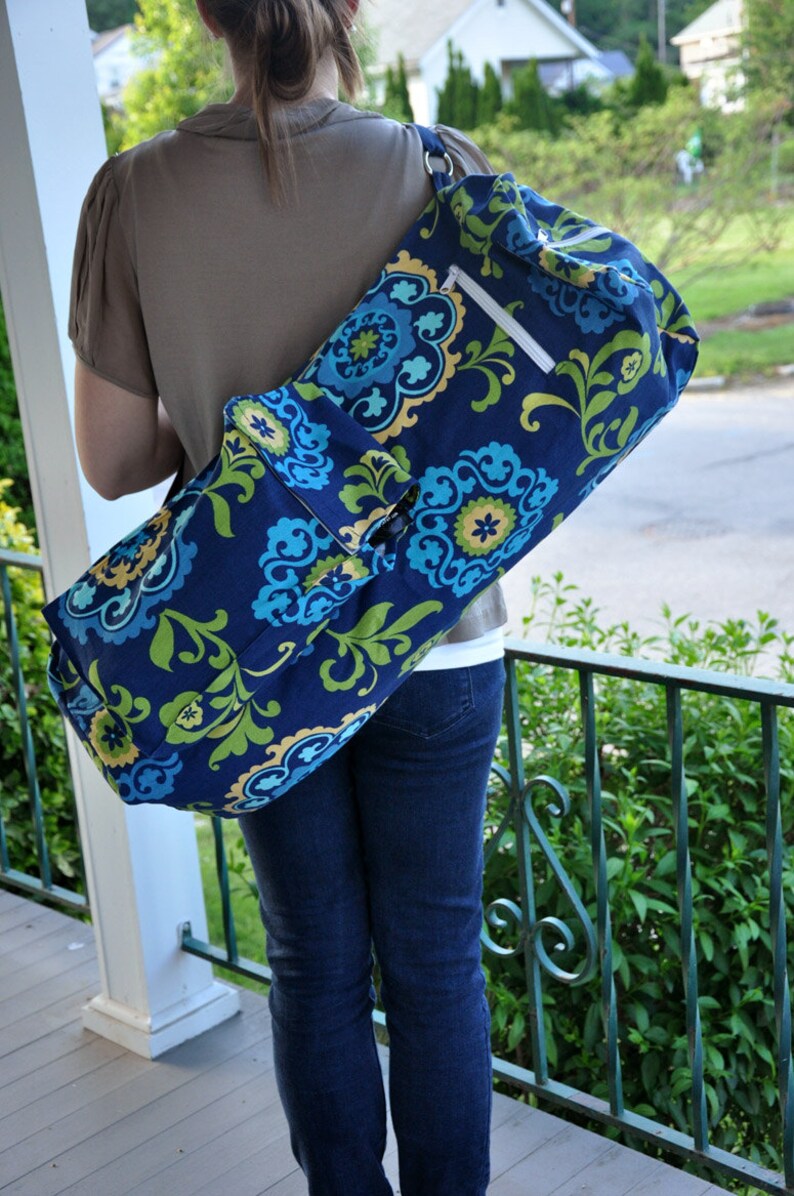 Pu&ograve; includere: Una donna porta una borsa per tappetino da yoga blu e verde con un motivo floreale e una tasca con cerniera. La borsa &egrave; indossata sulla spalla e ha una tracolla.