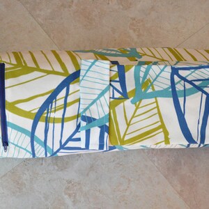 May include: A white yoga mat bag with a blue and green leaf pattern. The bag has a zipper closure and a small pocket on the side. The bag is rolled up and ready to be used.