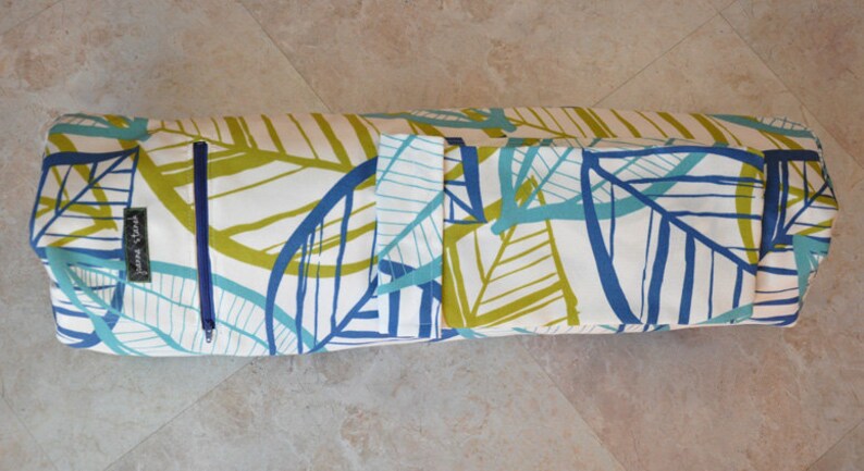 May include: A white yoga mat bag with a blue and green leaf pattern. The bag has a zipper closure and a small pocket on the side. The bag is rolled up and ready to be used.