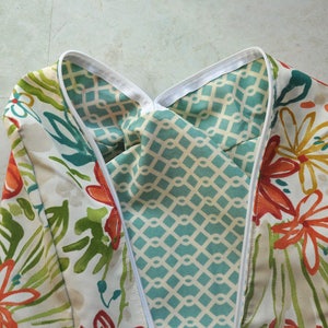 May include: A white fabric with a floral pattern in shades of orange, green, and teal. The fabric is folded in half, revealing a turquoise and white geometric pattern on the inside.