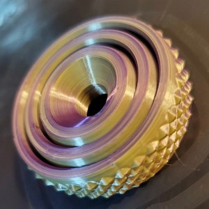 May include: A 3D printed object with a layered, concentric design. The object is a light gold color with a purple hue in the center. The outer edge of the object has a textured, diamond-shaped pattern.