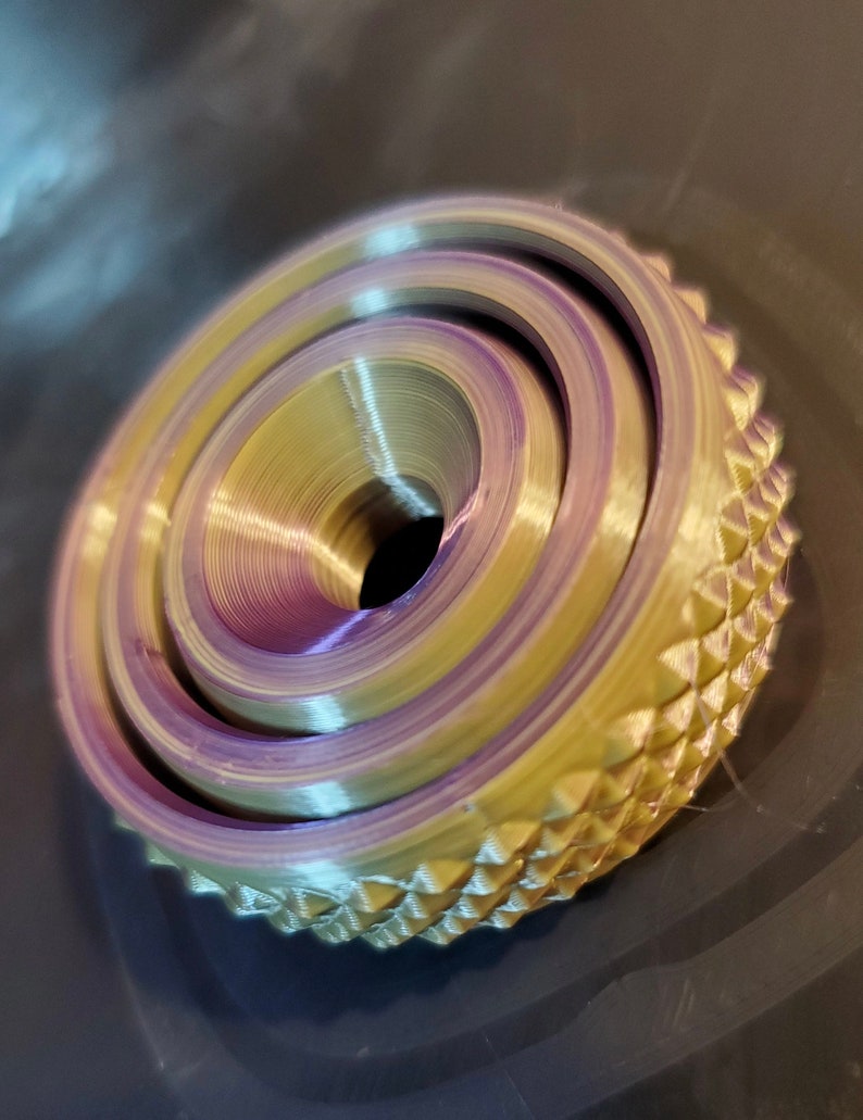 May include: A 3D printed object with a layered, concentric design. The object is a light gold color with a purple hue in the center. The outer edge of the object has a textured, diamond-shaped pattern.