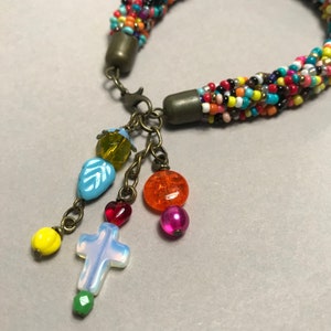 May include: A colorful beaded bracelet with a variety of charms. The bracelet features a braided design with small, multicolored beads. Charms include a cross, hearts, and various colorful beads. The bracelet is secured with a clasp.
