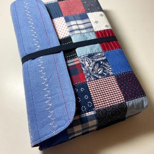 May include: A patchwork fabric case with a blue denim flap and a black elastic band. The patchwork features squares of red, blue, and white patterned fabrics. White stitching decorates the flap.