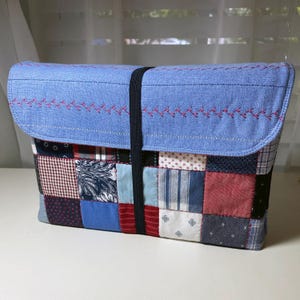 May include: A patchwork fabric case with a light blue flap, secured by a black elastic band. The flap features pink embroidered detailing. The body of the case is composed of various fabric squares in red, blue, and white patterns.