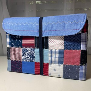 May include: A patchwork laptop sleeve featuring a variety of fabric patterns in blue, red, and white. The top flap is light blue with white zigzag stitching. A dark blue elastic band secures the sleeve.