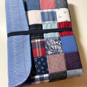 May include: A patchwork journal cover with a light blue flap featuring decorative stitching. The cover is made of various fabric squares in shades of blue, red, and white, held together by a black elastic band.