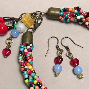 May include: A colorful beaded necklace and matching earrings set. The necklace is made of multiple strands of small, multicolored beads. The earrings feature red heart-shaped beads and light blue beads. The set includes charms and a star-shaped bead.
