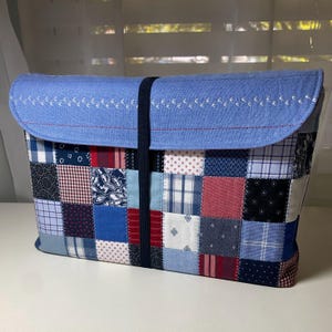 May include: A patchwork laptop sleeve featuring a variety of blue, red, and white fabric squares. The flap is light blue with white embroidered birds and a red stitched border. A black elastic band secures the flap.