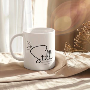 May include: White ceramic mug with the words "Be Still" in a black script font. The mug also includes the text "and know that I am God" and "Psalms 46:10". The mug is on a beige surface.