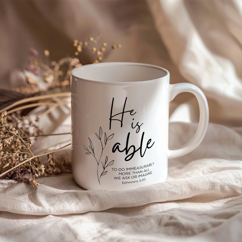 K&ouml;nnte beinhalten: Wei&szlig;e Keramiktasse mit einem schwarzen Design, das "He is able" sagt, mit einem kleinen Blumenmuster und dem Text "To do immeasurably more than all we ask or imagine Ephesians 3:20"