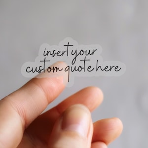 Puede incluir: Una pegatina transparente con el texto "insert your custom quote here" en un estilo manuscrito. La pegatina se sujeta entre dos dedos sobre un fondo gris borroso. La pegatina tiene forma de nube.