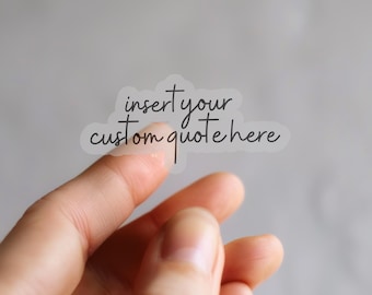 Op de afbeelding: Een transparante sticker met de tekst "insert your custom quote here" in een handschriftstijl. De sticker wordt tussen twee vingers gehouden tegen een vage grijze achtergrond. De sticker heeft de vorm van een wolk.