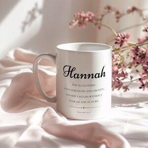 Könnte beinhalten: Eine weiße Keramiktasse mit schwarzem Henkel. Auf der Tasse steht der Name "Hannah" sowie das Zitat "Sie ist bekleidet mit Stärke und Würde, und sie lacht ohne Angst vor der Zukunft." Das Zitat stammt aus Sprichwörter 31:25.