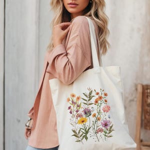 Op de afbeelding: Een witte canvas tote bag met een bloemenprint met verschillende bloemen in tinten oranje, geel, roze en paars. De bloemen zijn in een cluster gerangschikt en omgeven door groene bladeren.
