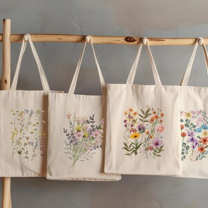 Op de afbeelding: Vier natuurlijke canvas tassen met verschillende bloemenontwerpen. Elke tas heeft een boeket kleurrijke wilde bloemen, waaronder roze, paarse, gele en oranje bloemen.