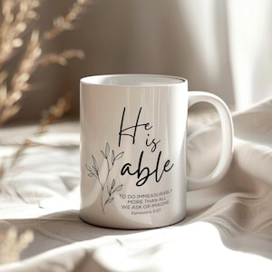 K&ouml;nnte beinhalten: Eine wei&szlig;e Keramiktasse mit schwarzem Text, der "He is able" lautet, und einem kleinen Blumenmuster. Der Text setzt sich fort mit "To do immeasurably more than all we ask or imagine, Ephesians 3:20."