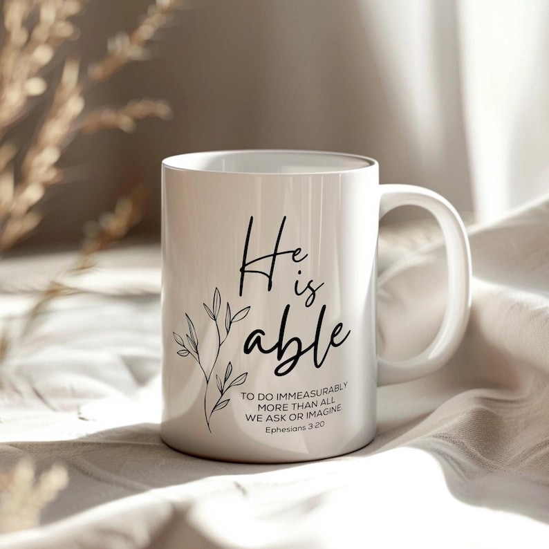 K&ouml;nnte beinhalten: Eine wei&szlig;e Keramiktasse mit schwarzem Text, der "He is able" lautet, und einem kleinen Blumenmuster. Der Text setzt sich fort mit "To do immeasurably more than all we ask or imagine, Ephesians 3:20."