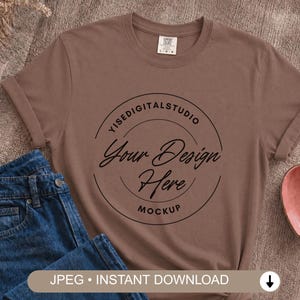 Puede incluir: Una camiseta marrón de cuello redondo con un gráfico circular negro con el texto YISEDIGITALSTUDIO, Your Design Here y MOCKUP. La camiseta se combina con pantalones vaqueros azules. La etiqueta muestra el texto Comfort Colors y la talla L-G-G.
