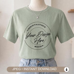 Puede incluir: Una camiseta de manga corta en color verde salvia con un gráfico circular negro en el pecho que incluye el texto YISEDIGITALSTUDIO, Your Design Here y MOCKUP. La prenda se combina con unos vaqueros azul claro y un collar plateado.