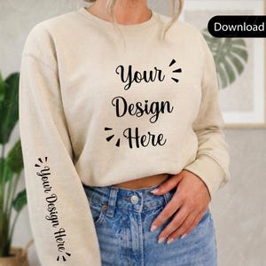 Puede incluir: Una sudadera beige con el texto "Your Design Here" en escritura negra en la parte delantera y la manga. La modelo lleva vaqueros azules. La imagen es una maqueta para un diseño de ropa.