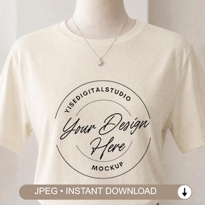 Puede incluir: Una camiseta de manga corta de color crema con un gráfico circular negro en el pecho. El gráfico contiene el texto YISEDIGITALSTUDIO, Your Design Here y MOCKUP. Un delicado collar de plata con un pequeño colgante se lleva al cuello.