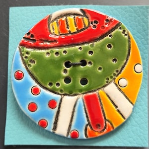 May include: A large, colorful ceramic button with a unique design. The button features a central green section with black buttonholes, surrounded by red, orange, blue, and white sections. Red dots and white stripes add detail. The button is on a light blue background.