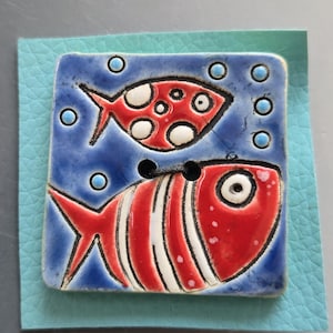 May include: A square ceramic button with a blue background and two red fish designs. The top fish has white spots, and the bottom fish has white stripes. The button is mounted on a light blue leather square.