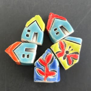 May include: A collection of five ceramic house-shaped ornaments. Each miniature house features a unique color scheme, including blue, red, yellow, and green, with hand-painted details like windows, doors, and a butterfly or leaf design. The ornaments are arranged in a circular pattern.