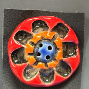 May include: A ceramic flower-shaped object with a glossy finish. The flower has a red outer ring, orange petals, and a blue center with black accents. It is set on a textured black square.