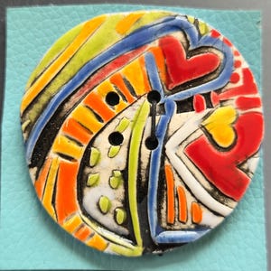 May include: A colorful, ceramic button with a vibrant abstract design. The button features a mix of red, yellow, orange, blue, and green hues, with black accents and three holes for sewing. It is set on a square of light blue leather.