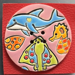 May include: A round, ceramic button with a pink background featuring a blue dolphin with a yellow heart, an orange seahorse, and a yellow, green, and blue sea creature. The button is attached to a red leather square.