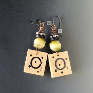May include: Handmade earrings with light wood square tiles, each with a black circle and the number 1. Olive green and black beads, copper and silver accents, and black ear wires complete the design.