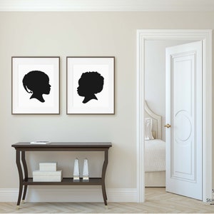 May include: Two black and white silhouette portraits of children framed in simple black frames, hanging on a white wall above a dark brown console table with a white box and two silver candle holders on the top shelf.